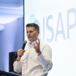 Prof. Kelly Swanson holding a microphone and speaking in front of a screen that shows the ISAPP logo.