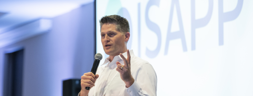 Prof. Kelly Swanson holding a microphone and speaking in front of a screen that shows the ISAPP logo.