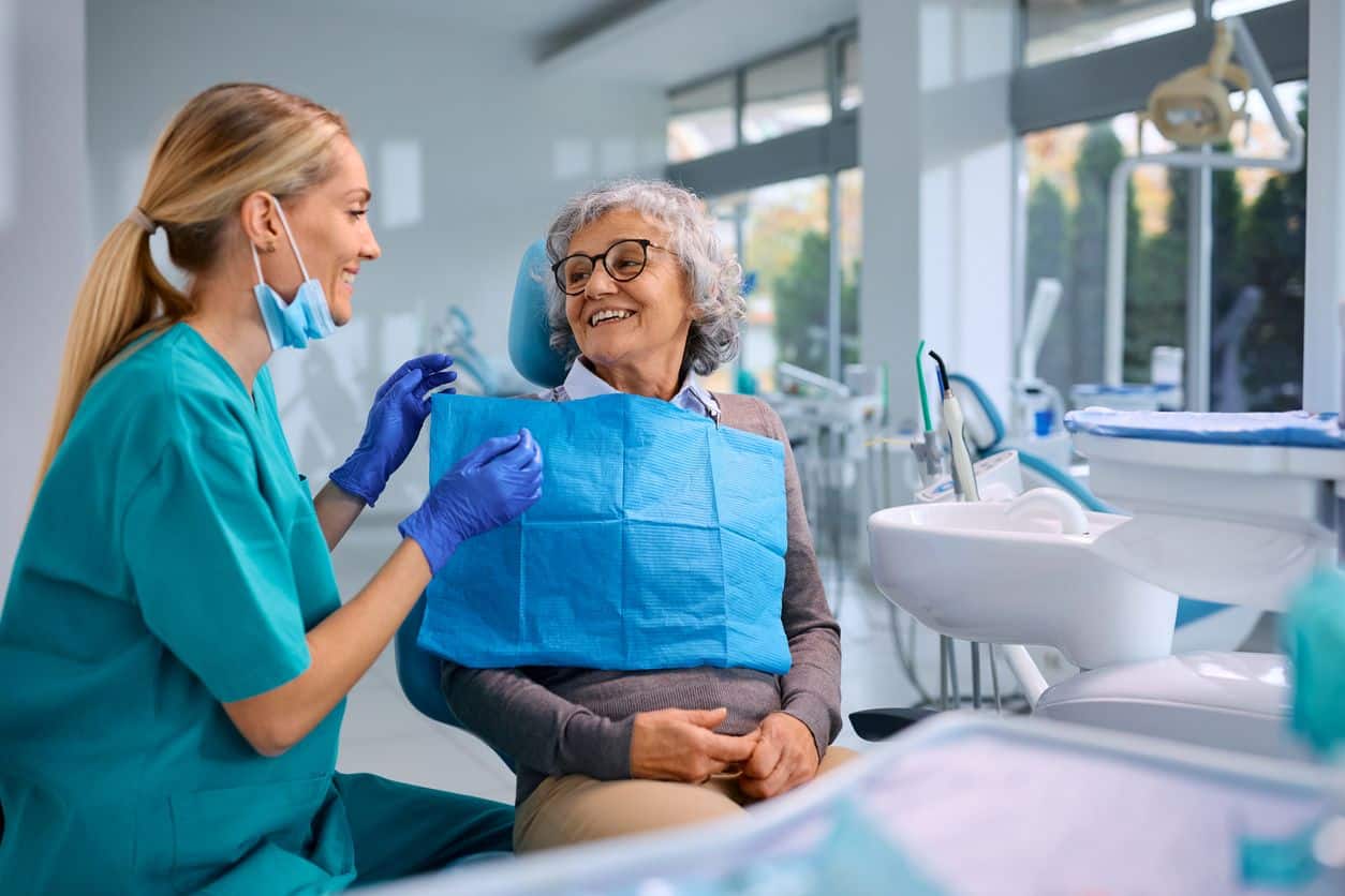 Dentist sitting beside older woman in the patient chair of a dental office, adjusting a bib around her neck.