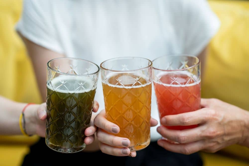 Three glasses of kombucha (green, yellow and pink) being held together with blurry people in background.